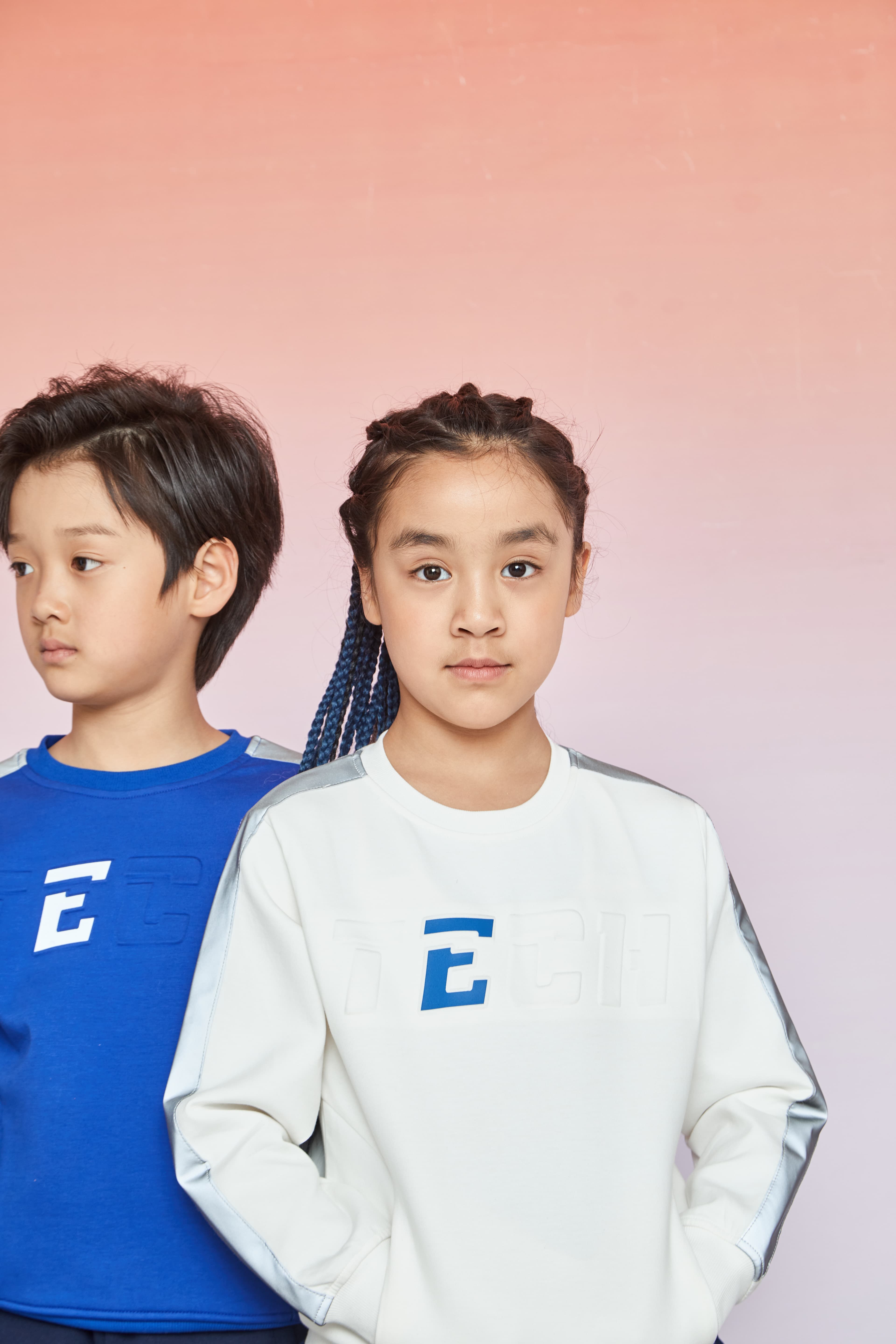 Girl with blue braids and boy in branded sweatshirts pose against a pink background.