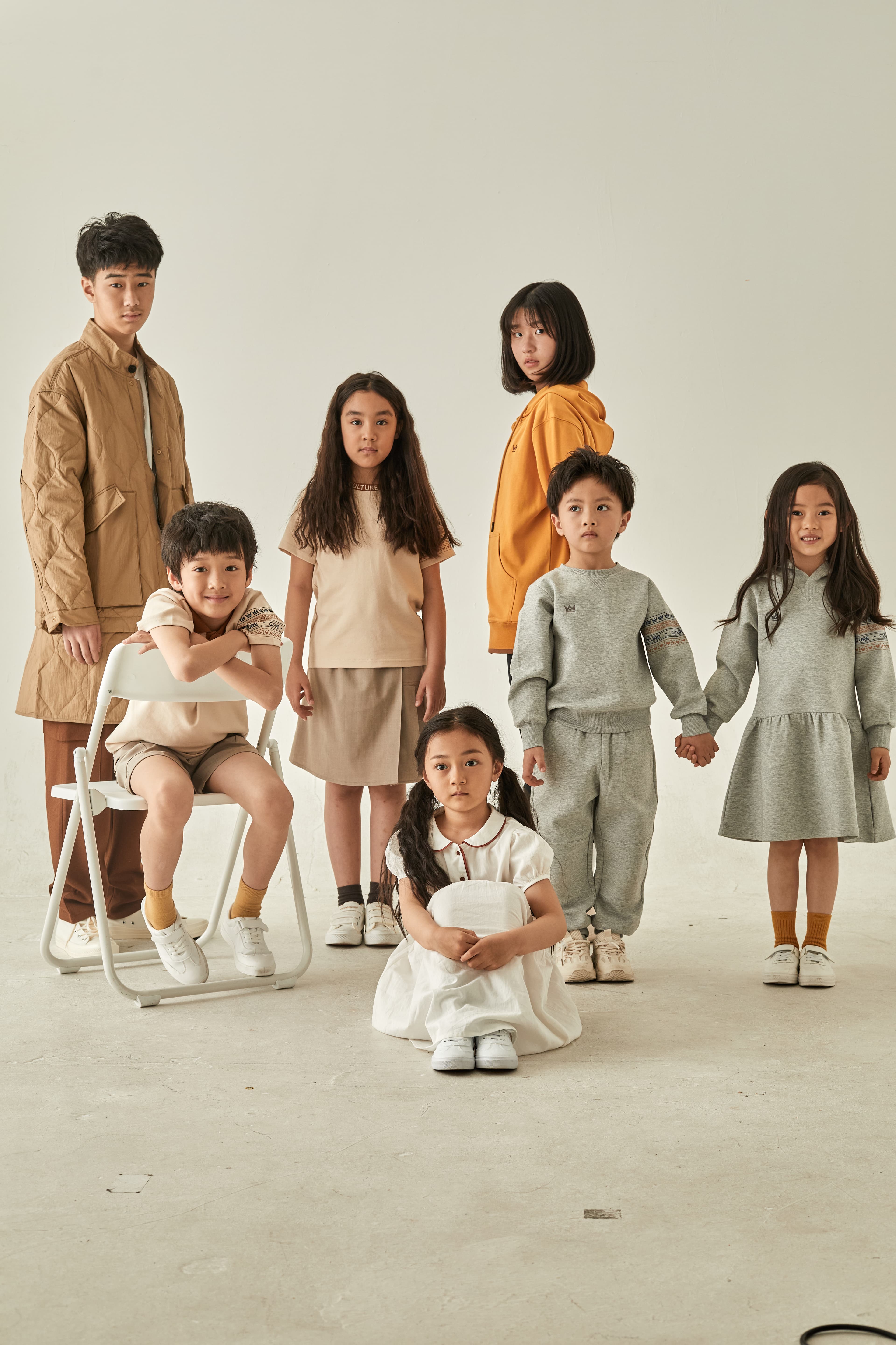 Seven children in beige, grey, and orange clothing pose in a minimalist white studio.