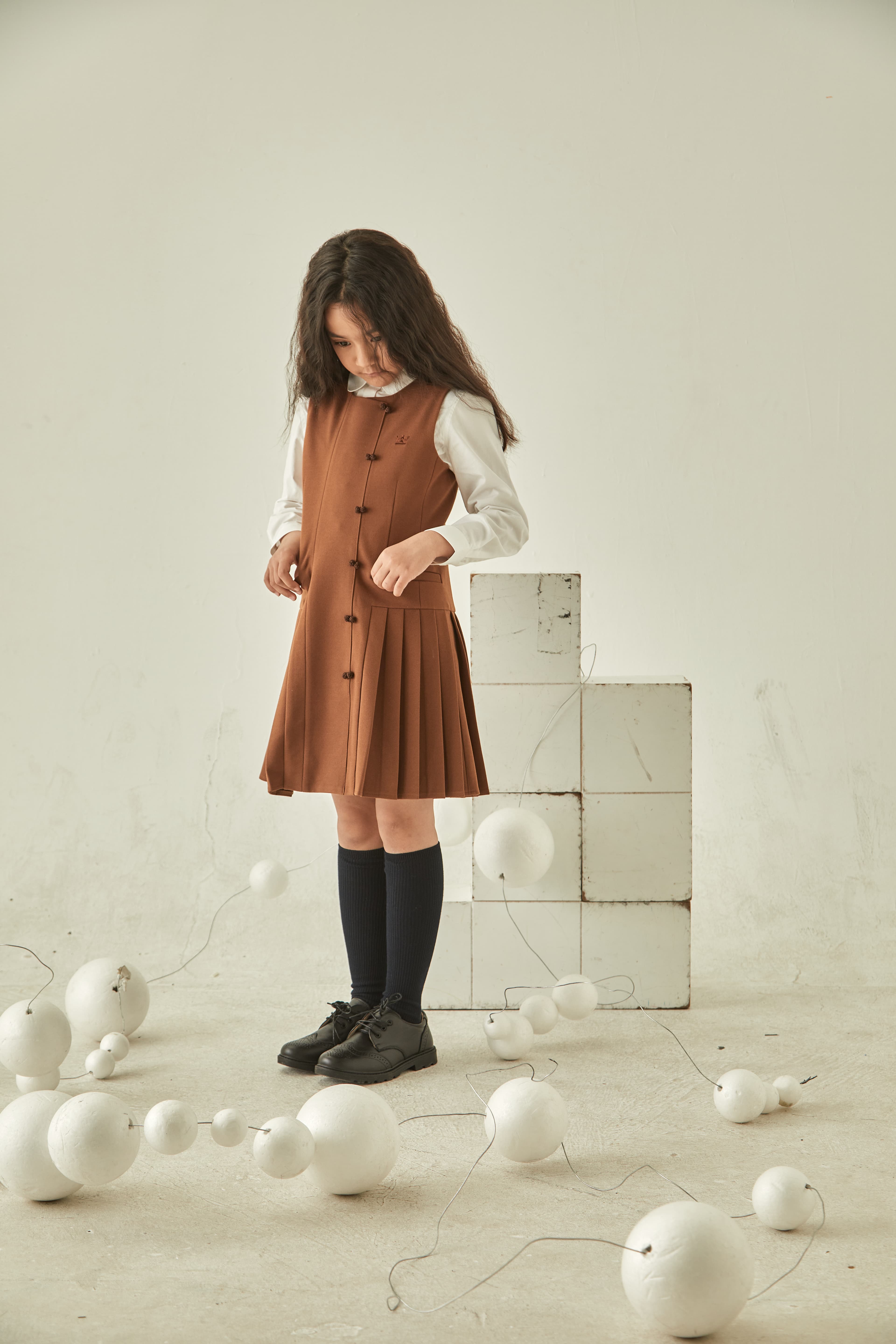 Girl in brown pleated dress looking down at white spheres in a minimalist studio.