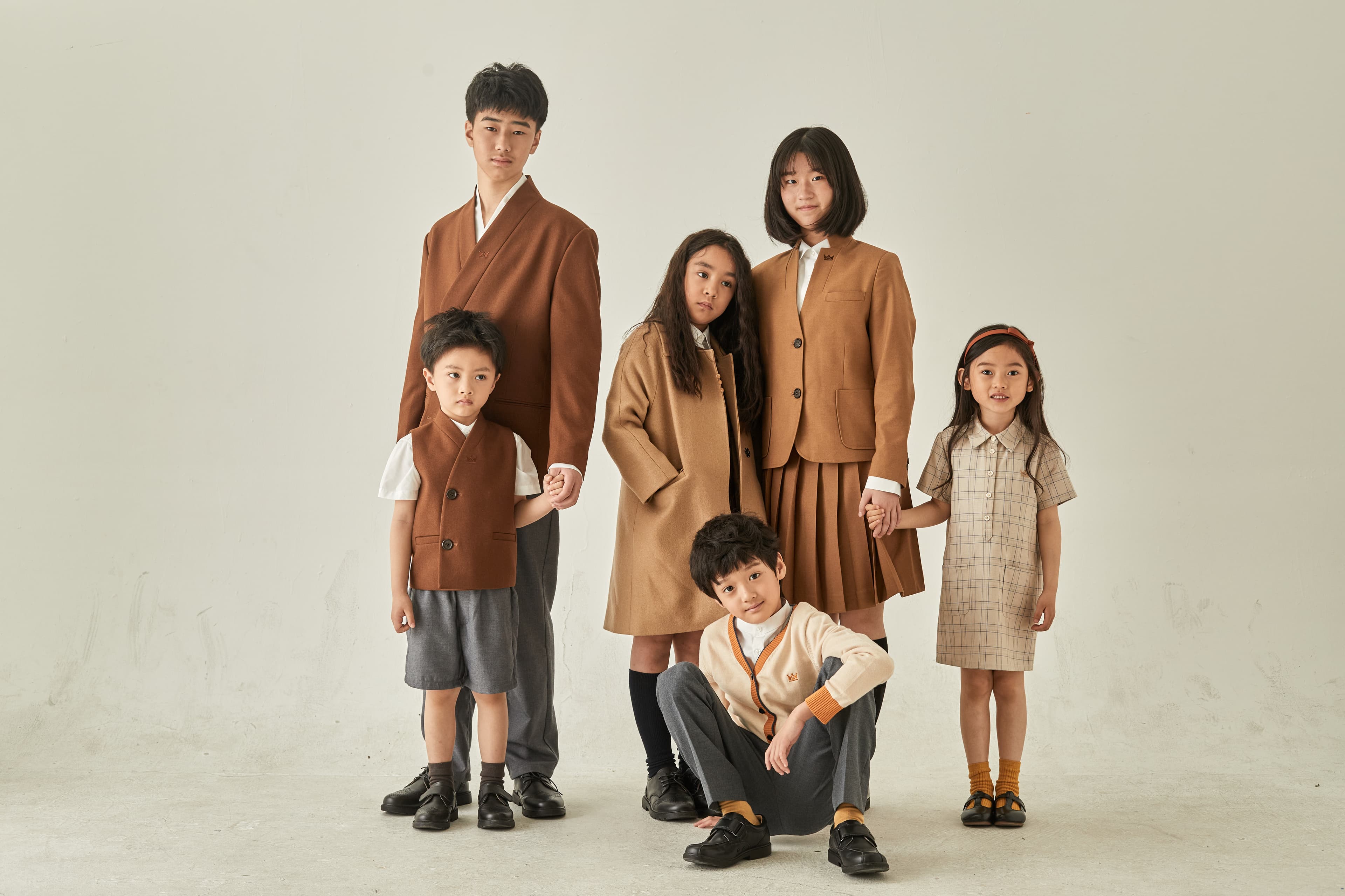 Six children in coordinated brown and tan outfits pose against a plain, light-colored background.