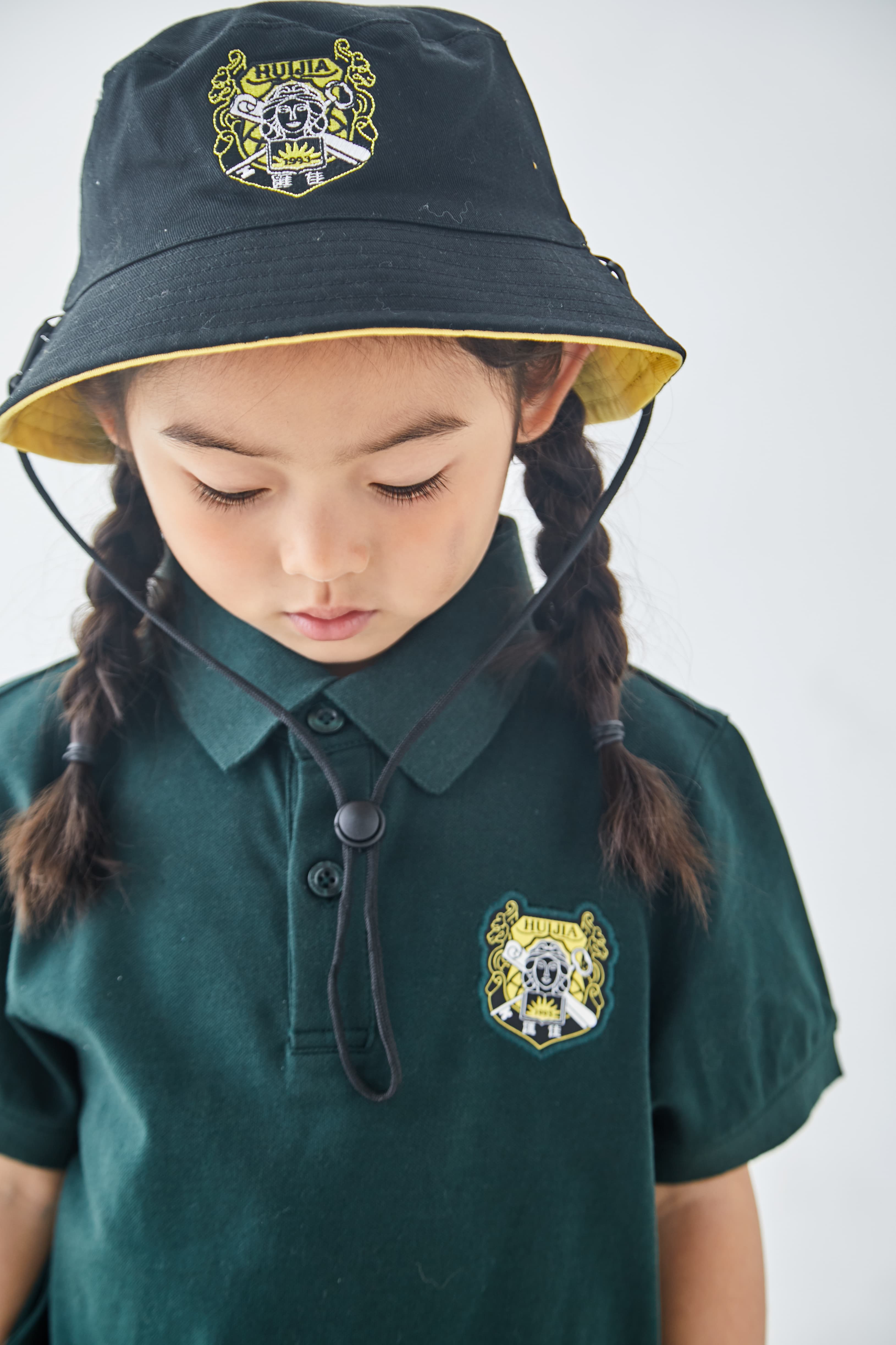 Young girl with braids looking down, wearing a green school uniform and black bucket hat.